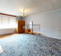 The interior of a cabin with a wooden wall, a tiled stove, and simple furniture in a cozy room.