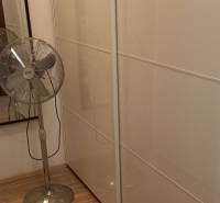A wardrobe in a 2-room apartment with white sliding wardrobes and a standing fan.