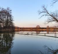 A lake with a small bridge, surrounded by bare trees and in the background the warm colors of a sunset.