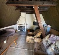 The interior of a cabin with older furniture and a fireplace, wooden beams on the ceiling.