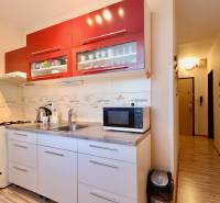 A kitchen space in a studio apartment with a white kitchen unit, red cabinets, and a wooden-patterned floor.