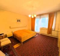 Spálňa v rodinnom dome s kobercovou podlahou, veľkým oknom a starším nábytkom.