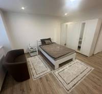 A room with a bed, a chair, and a wardrobe in a studio apartment, with a wooden decor floor.