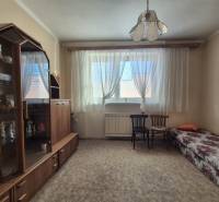 A room in a family house with a display cabinet, bed, chairs, and carpet, illuminated by a window with curtains.