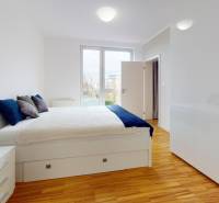 A bright bedroom with a bed, wood-patterned flooring, and a large window in a two-room apartment.