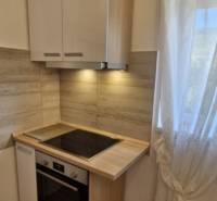 A kitchenette with a cooktop, white cabinets, and a window; flooring with a wood design in a 2-room apartment.