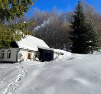 A snow-covered cottage in Píla surrounded by winter nature, smoke rising from the chimney.