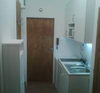 A kitchenette in a one-room apartment with white cabinets and wooden doors.