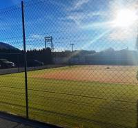 A tennis court by a family house in Necpaly, fenced and illuminated by sunlight.