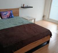 A bedroom in a one-room apartment with a wood-patterned floor, a bed, and an office chair.