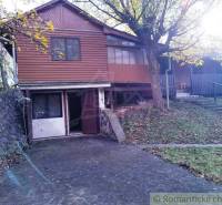 A cottage in Dudince with a stone wall and wooden facade surrounded by trees.