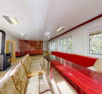 A room in a cottage with a stove, sofas, wooden tables, and a view of the forest.