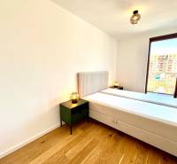 A bright bedroom in a two-room apartment with a wood-patterned floor and a large window.