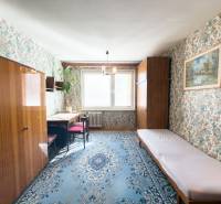 A room in a 3-room apartment with floral wallpaper, a blue carpet, and furniture with a wood finish.