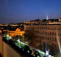 An evening view from the balcony of a 3-room apartment overlooking the illuminated city with buildings and greenery.