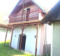 A red balcony on a cottage in Hontianske Trsťany, next to a green lawn and a neighboring house.