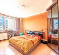 A bedroom with an orange wall, a bed, and a wooden decor floor in a three-room apartment.