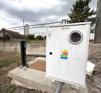An electricity meter on a concrete pedestal by a family house in Komárno under a cloudy sky.