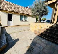 A family house with a sloped roof, a paved yard, and greenery, surrounded by a clear sky.