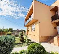 A family house in Okoč with a front garden and landscaped greenery in sunny weather.