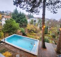 A wooden terrace with a pool surrounded by greenery and a view of houses in Bratislava.