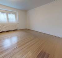 A room with white walls, a large window, and a wooden-patterned floor in a family house.