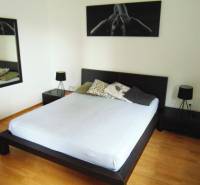 Bedroom of a one-room apartment with a double bed, mirror, and wooden decor flooring.