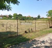 A flat plot of land bordered by a fence in the Gardens of Marcelová, with houses visible in the background.