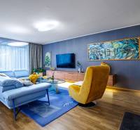 A living room in a family house with a blue sofa, a yellow armchair, and a wooden-patterned floor.