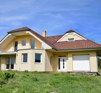 A family house in Buková with a sloped roof, light facade, and garage, surrounded by greenery.