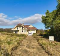 A family house in Buková with an access road, a car, and surrounding nature under a blue sky.
