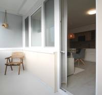 A furnished balcony with a wooden chair leads into a studio apartment through glass doors.