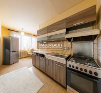 The kitchen of a three-room apartment with dark cabinets, light walls, and a large window.