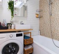 Bathroom in a 2-room apartment with a washing machine, bathtub, and shelves, white tiles.