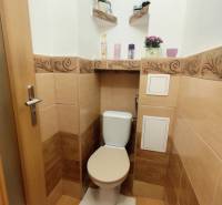 A toilet in a 3-room apartment with beige tiles and decorative shelves.