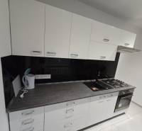 A kitchen in a 2-room apartment with white cabinets and appliances, and a wooden decor floor.