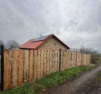 A cottage in Šahy with a red roof and a wooden fence by a country road.