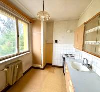 A kitchen in a 2-room apartment with a large window, older furniture, and a light wall.
