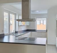 A kitchen in a family house with large windows, a stove in the middle, and light-colored furniture.