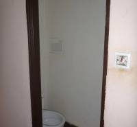 A toilet in a small two-room apartment with plain walls and ceramic tiles on the floor.