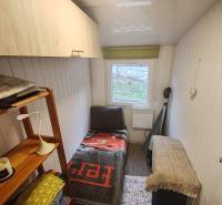 A small room in a cabin with a bed, a shelf, and a window, wooden cabinets and walls.