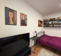 A studio apartment with a bed, bookshelves, and a television, with a wood-patterned floor.