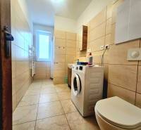 A bathroom with a washing machine and toilet in a 2-room apartment, with ceramic tiles and a bright window.