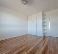 A room in a 2-room apartment with a wood-patterned floor, a white wardrobe, and shelves.