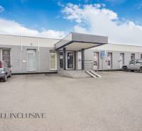 A single-story building with commercial spaces on Vajanského Street, a parking lot, and cars in front of the entrance.