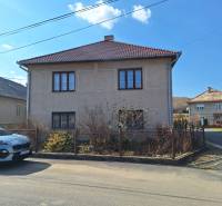 A family house in Tachtoch with a fence, a car, and a neighboring house under a blue sky.