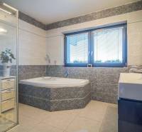 Bathroom in a family house with a bathtub, shower, and sink, light tiles, window with blinds.
