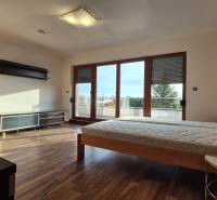 A bedroom in a family house with a wooden decor floor, a bed, and large balcony windows.