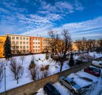 Snow-covered yard with parking lot, building, and trees on Strojárenská Street in Košice.