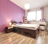 A bedroom with a light purple wall, a bed, and a wooden decor floor in a 3-room apartment.